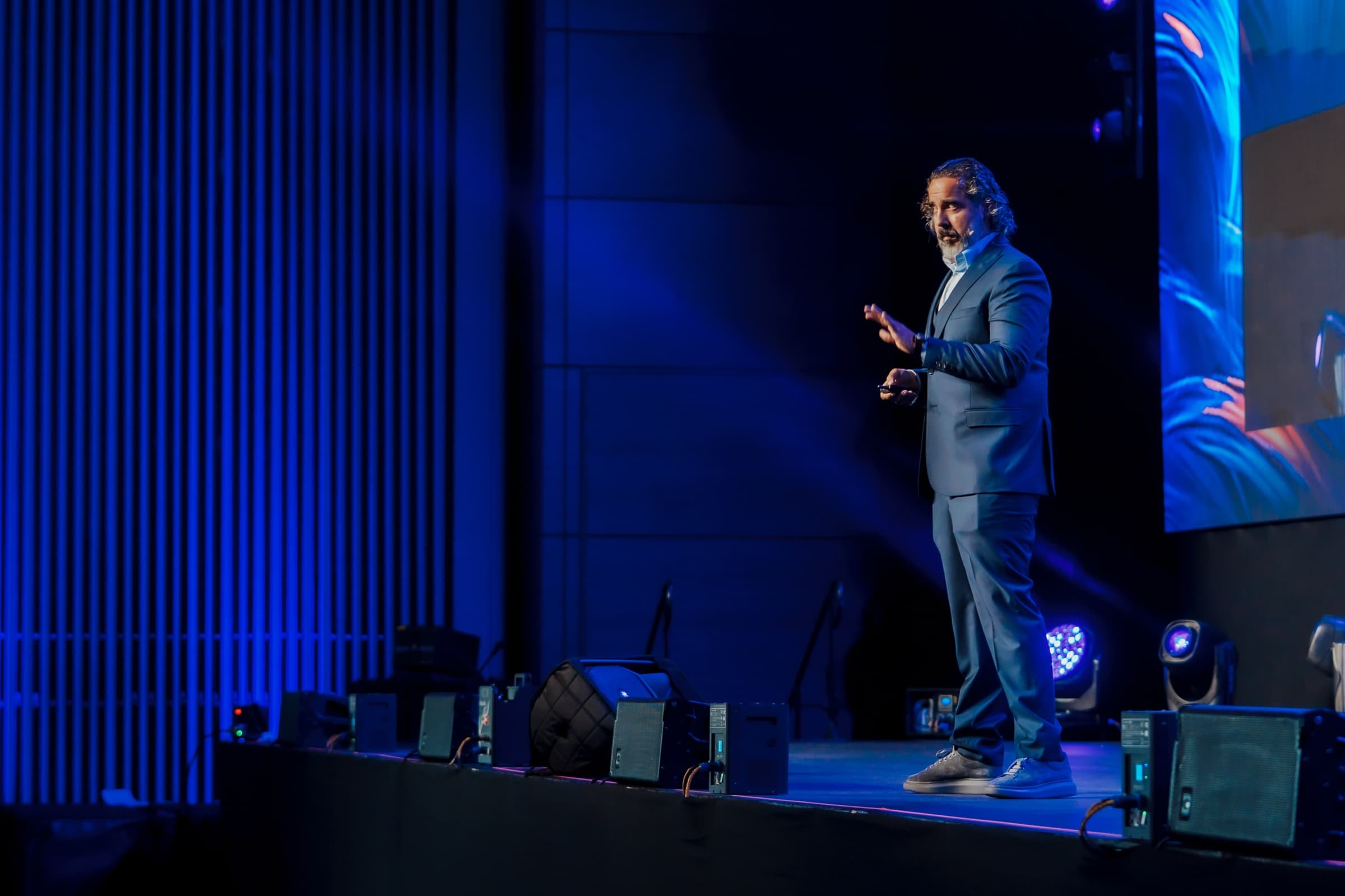 Manuel Suarez alone in a spotlight on the Game Changers stage in Bogotá, mid-sentence, one hand raised, the stage rig glowing behind him