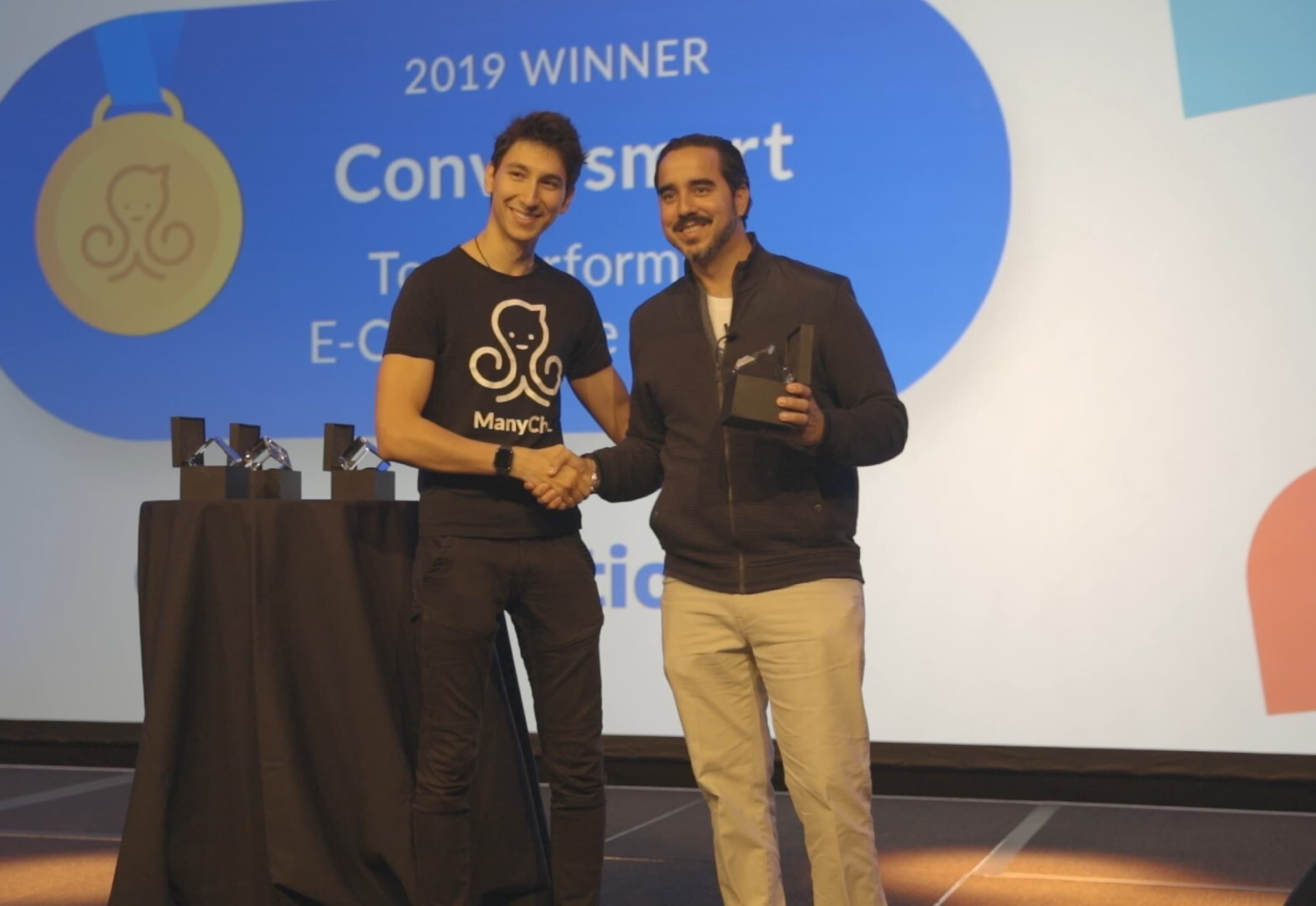 Manuel Suarez accepts a glass award on stage at the ManyChat Conversations conference, a row of trophies on a draped table beside him.