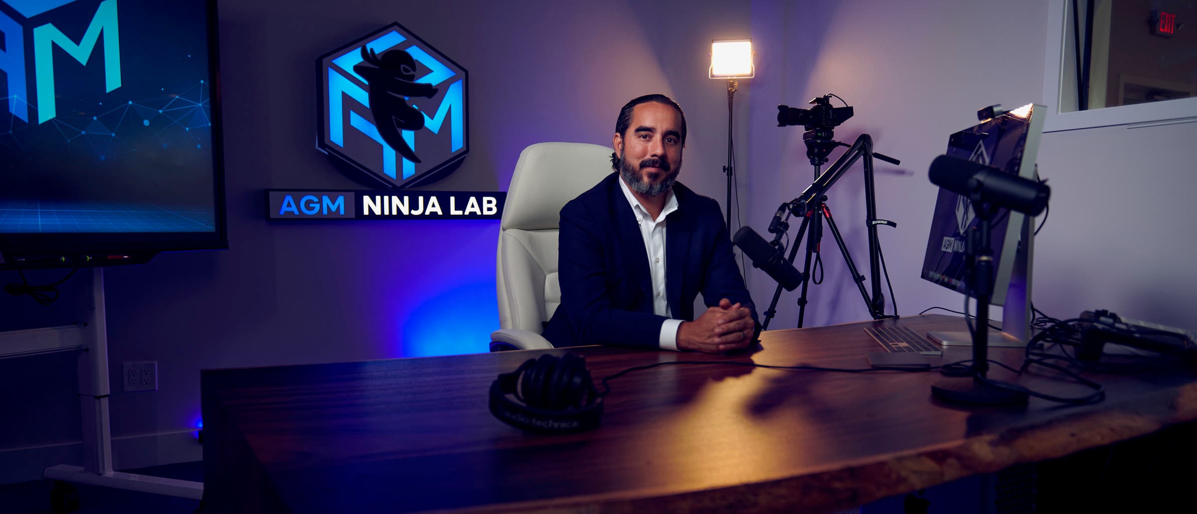 Manuel Suarez at the desk in the AGM Ninja Lab studio, the illuminated ninja mark and the AGM Ninja Lab sign on the wall behind him, camera and microphone rigged around the desk