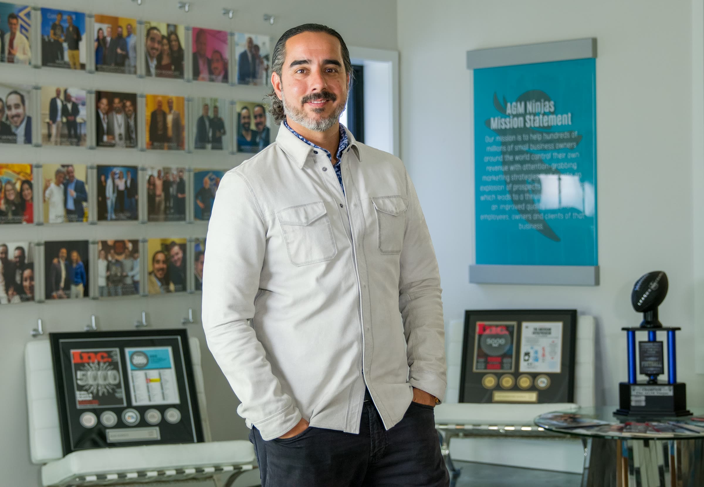 Manuel Suarez standing in the AGM office between the wall of framed photographs and the AGM Ninjas mission statement panel, which carries a ninja mask silhouette