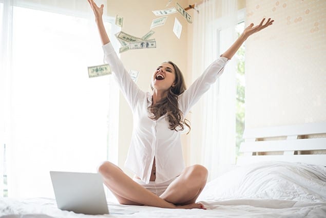 happy woman sitting in a bed with laptop and throwing money